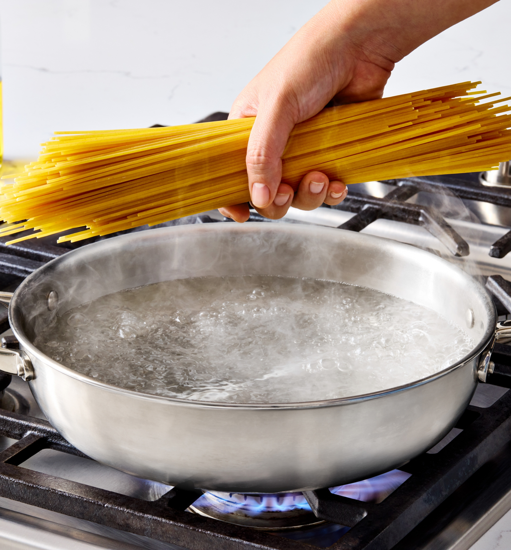 Hand adding dry spaghetti to a pan of boiling water on a gas stove